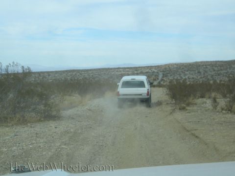1990 Ford Bronco - white SUV kicks up dust driving away on a flat desert two-track trail with scrub brush and distant hills.