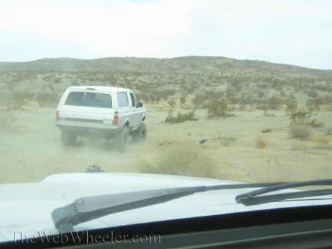 1990 Ford Bronco - View through a vehicle windshield of a white SUV raising dust on a desert trail, with the wiper and hood visible in frame.