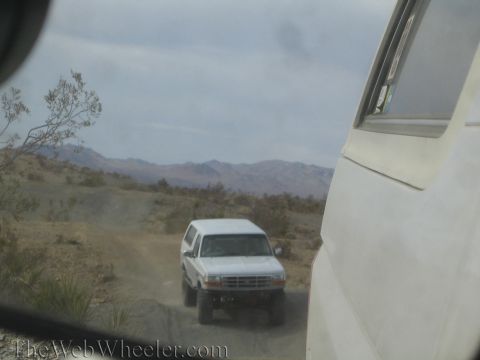 1990 Ford Bronco - white Ford SUV with a brush guard drives a desert trail toward distant mountains, framed past the body of a parked vehicle.