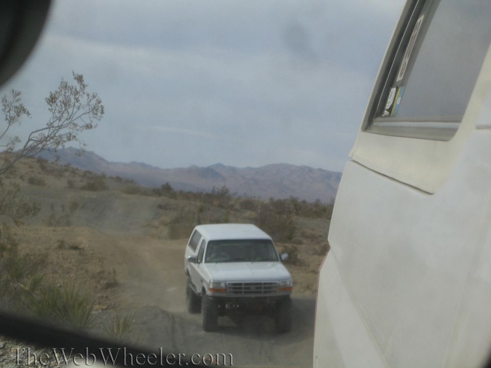 White Ford SUV on a desert trail with mountains, seen past a parked vehicle