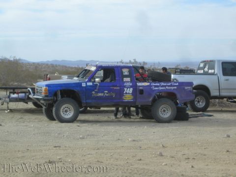 1990 Ford Bronco - blue prerunner-style off-road race truck marked 748 and BFGoodrich parked in the desert beside support trucks and a trailer.