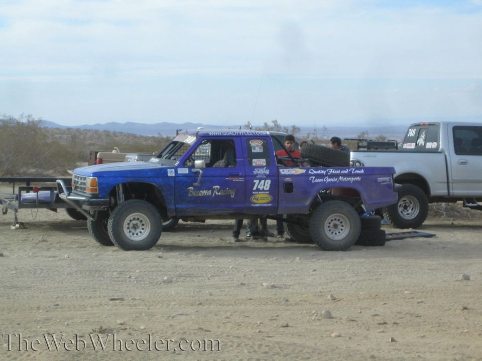 Blue off-road race truck number 748 parked in the desert with chase trucks
