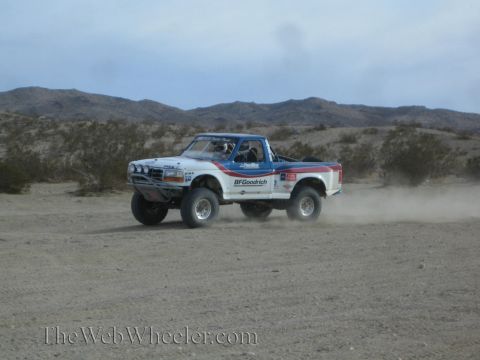 1990 Ford Bronco - white, blue and red Ford prerunner race truck with BFGoodrich graphics kicks up dust racing across a desert flat below mountains.