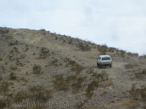 1990 Ford Bronco - white SUV climbs a steep, loose rocky desert hillside dotted with scrub under a gray overcast sky.