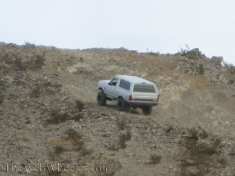 1990 Ford Bronco - light gray SUV with a roof rack crests a steep, rocky desert ridge during an off-road hillclimb.