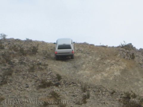 1990 Ford Bronco - silver SUV works up a steep, loose rock-strewn slope under a hazy overcast sky, viewed from downhill.