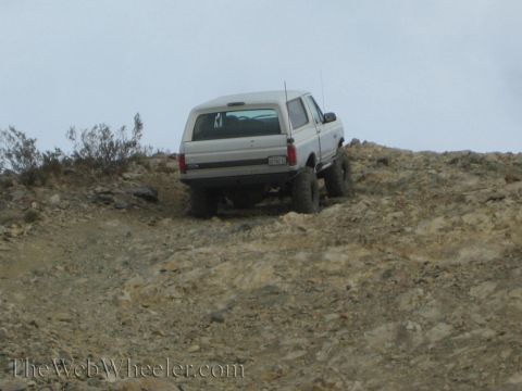 1990 Ford Bronco - lifted white SUV with a visible Ford tailgate badge climbs a steep, rocky desert slope on large off-road tires.