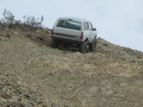 1990 Ford Bronco - lifted white Ford SUV with antenna and tailgate badge ascends a steep, rocky and dusty hillside under an overcast sky.