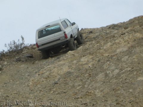 1990 Ford Bronco - lifted silver SUV tilts as it climbs a steep, rocky off-road slope, with off-road tires gripping loose rock.