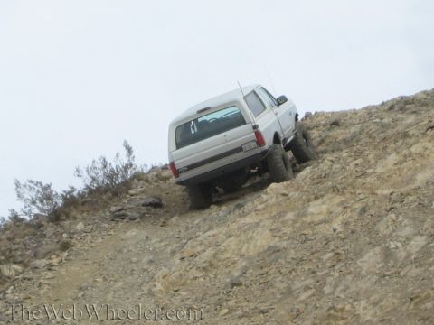 1990 Ford Bronco - lifted white SUV leans on a steep, loose rocky hillclimb under a flat gray sky, climbing on big off-road tires.