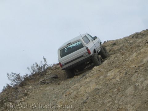 1990 Ford Bronco - lifted white SUV leans at a steep angle while crawling up a loose, rocky desert hillside under overcast skies.
