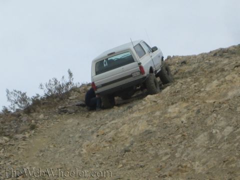 1990 Ford Bronco - lifted white SUV sits on a steep rocky slope while a person crouches near its rear wheel, apparently spotting or working on it.