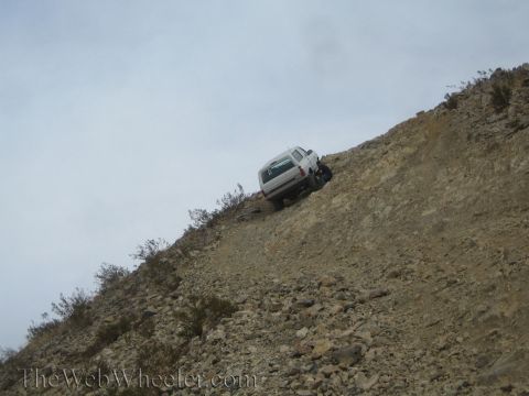 1990 Ford Bronco - distant silver SUV climbs a long, steep rock-covered desert slope under a pale overcast sky.