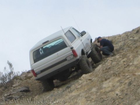 1990 Ford Bronco - lifted white Ford SUV is stopped on a steep rocky slope while a man in a blue shirt crouches by the rear wheel.