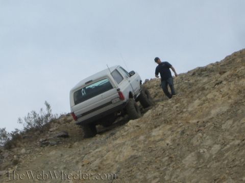 1990 Ford Bronco - silver lifted SUV crawls up a steep rocky desert grade while a man in a blue shirt stands on the slope nearby.