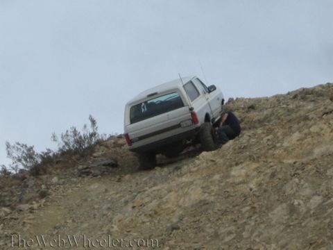 1990 Ford Bronco - silver lifted SUV sits angled on a steep dirt hillside as a crouching man inspects the rear wheel.