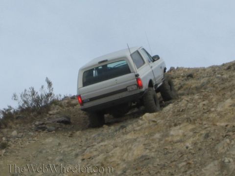 1990 Ford Bronco - Rear three-quarter view of a silver lifted SUV with oversized tires ascending a rocky desert hill.