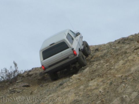 1990 Ford Bronco - silver lifted SUV leans at a steep angle on a rocky hillside with its tail lights glowing.