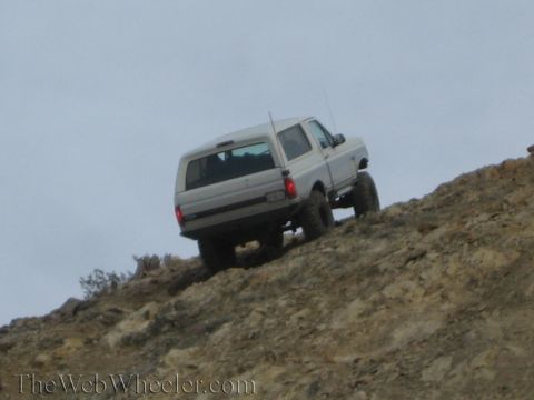 1990 Ford Bronco - silver lifted SUV with knobby tires works its way up a rocky off-road hill seen from behind.