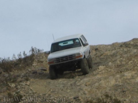 1990 Ford Bronco - white lifted Ford SUV climbs a rocky dirt hillside head-on, the Ford grille badge visible.