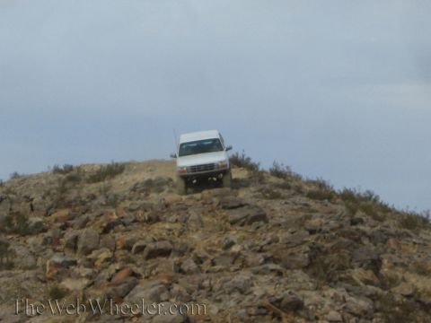 1990 Ford Bronco - white SUV crests a rugged rocky ridgeline in the distance under a cloudy desert sky.