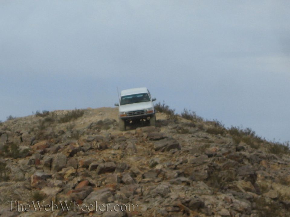White SUV cresting a distant rocky ridge under an overcast sky