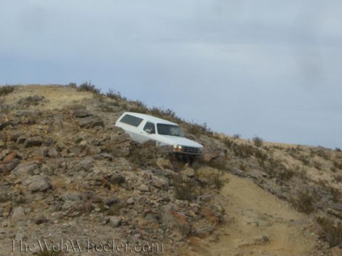 1990 Ford Bronco - white SUV navigates down a steep rocky desert slope along a narrow dirt trail.
