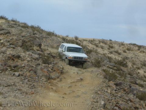1990 Ford Bronco - white SUV crawls up a rutted rocky desert trail flanked by scrub on a steep hillside.