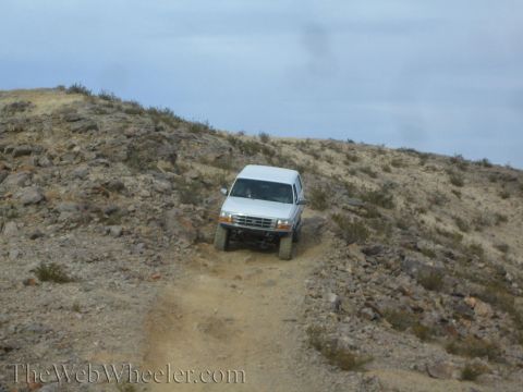 1990 Ford Bronco - white SUV climbs a sandy rutted trail flexing its suspension on a rocky desert hillside.