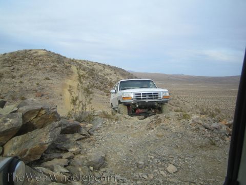 1990 Ford Bronco - white Ford truck sits on a rocky ridge trail overlooking a sweeping desert valley below.
