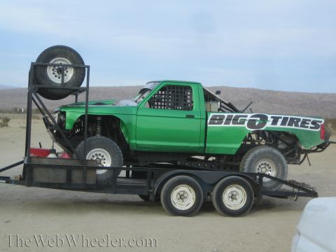 1990 Ford Bronco - green tube-chassis prerunner pickup with Big O Tires graphics sits strapped on a dual-axle trailer in the desert.