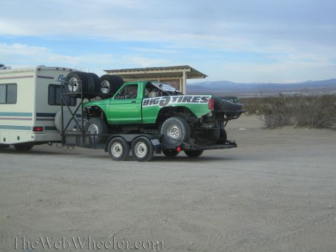 1990 Ford Bronco - green Big O Tires prerunner truck loaded on a trailer is parked behind a motorhome in a dusty desert lot.