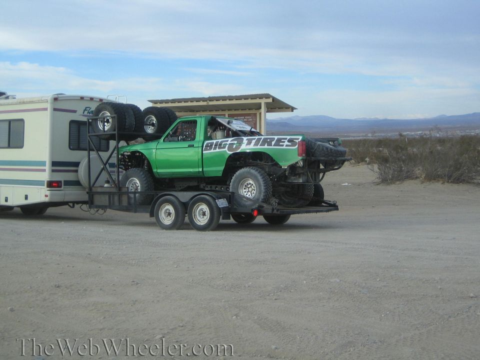 Green prerunner truck on a trailer behind an RV in a desert lot