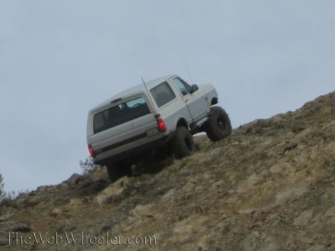 1990 Ford Bronco - silver lifted SUV with oversized tires crawls up a steep rocky desert hill, rear tail lights lit.