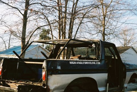 1978 Ford Bronco - A dark blue and gray Ford Bronco with its top removed, showing a bare roll cage and stripped interior, viewed from the rear three-quarter a…