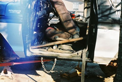 1978 Ford Bronco - Blue Bronco Custom mid-teardown outdoors, body up on jack stands with the door removed and set against a fender, exposing the bare seat fra…