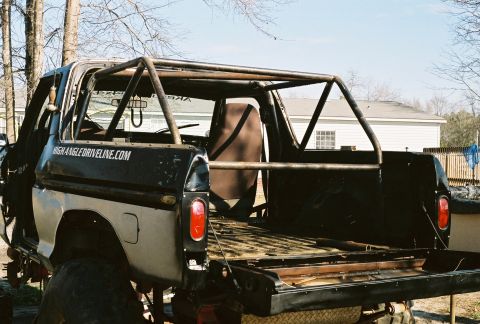 1978 Ford Bronco - Rear three-quarter view of a black Ford Bronco stripped down to a bare metal bed with a welded roll cage, a single brown bucket seat, and '…