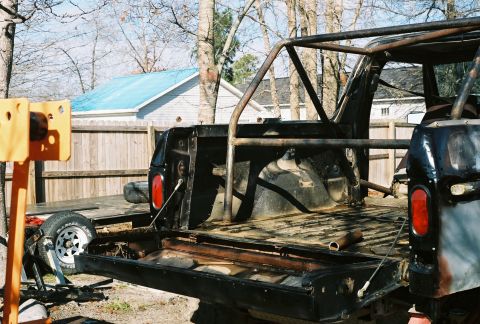1978 Ford Bronco - Stripped black full-size Bronco body in a backyard with the tailgate down and the roof removed, showing a bare welded roll cage, gutted rea…