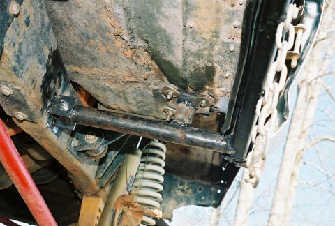 1978 Ford Bronco - Underbody view looking up at a rusty truck frame and floor pan, with a fabricated tubular link bolted to a welded bracket on the frame rail…