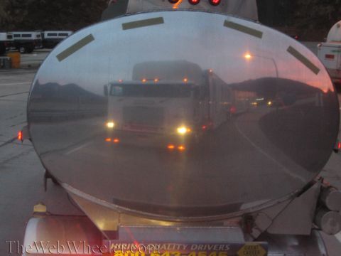1990 Ford Bronco - polished oval rear of a tanker trailer mirrors a semi truck with glowing headlights, with text on a placard below.