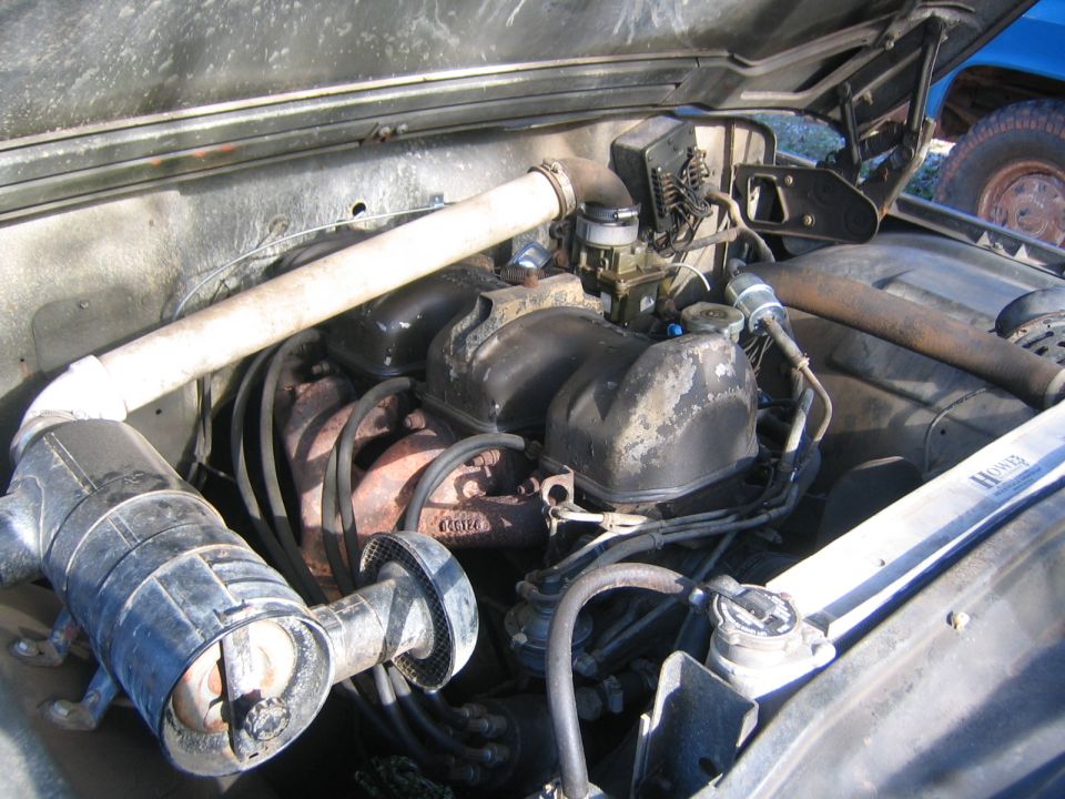 Engine bay with carburetor, air filter, and radiator visible. Another vehicle is partially seen in the background