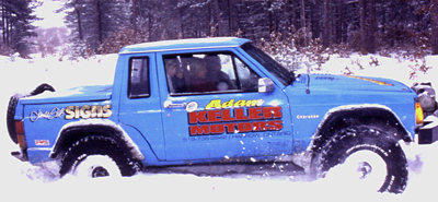 1982 Ford Bronco - A blue Jeep pickup truck navigates through a snowy landscape, kicking up snow as it goes.