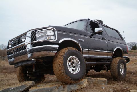 1995 Ford Bronco - This lifted 1995 Ford Bronco is shown off-road, navigating a rocky path with its large, mud-covered tires.
