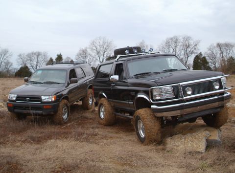 1995 Ford Bronco - Black lifted Ford Bronco with mud-packed tires parked beside a black Toyota 4Runner in a dry winter field, both shot from the front three-q…