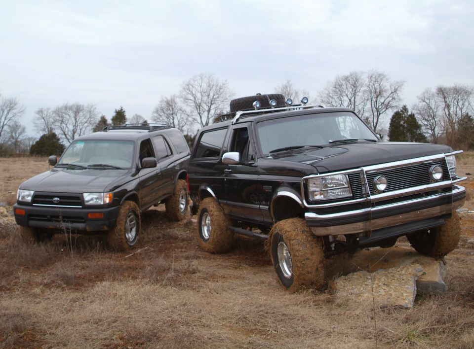 Muddy black lifted Ford Bronco parked next to black Toyota 4Runner in field