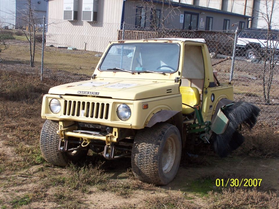 Yellow Suzuki Samurai with a large brush attachment on the rear, parked outdoors.