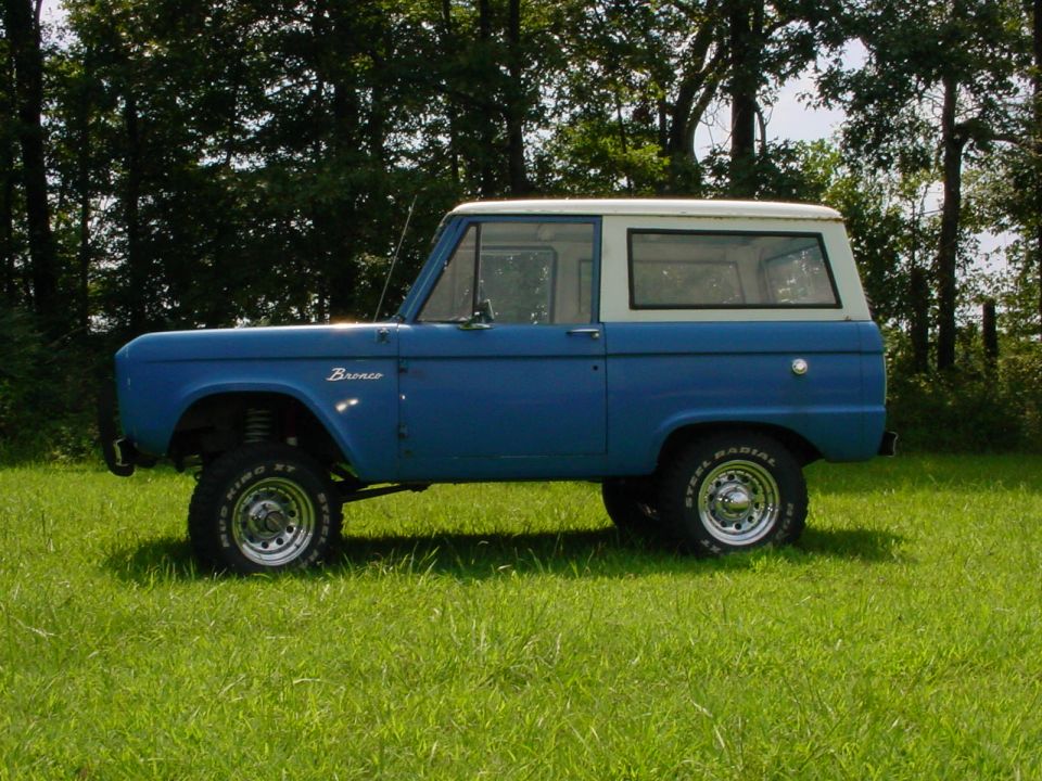 Blue and white classic Ford Bronco SUV in side view on grass with lifted suspension and off-road tires.