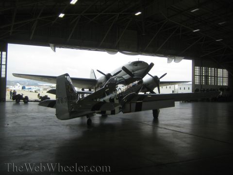 1990 Ford Bronco - silver fighter plane with black-and-white invasion stripes sits beneath the wing of a larger propeller aircraft in a hangar.