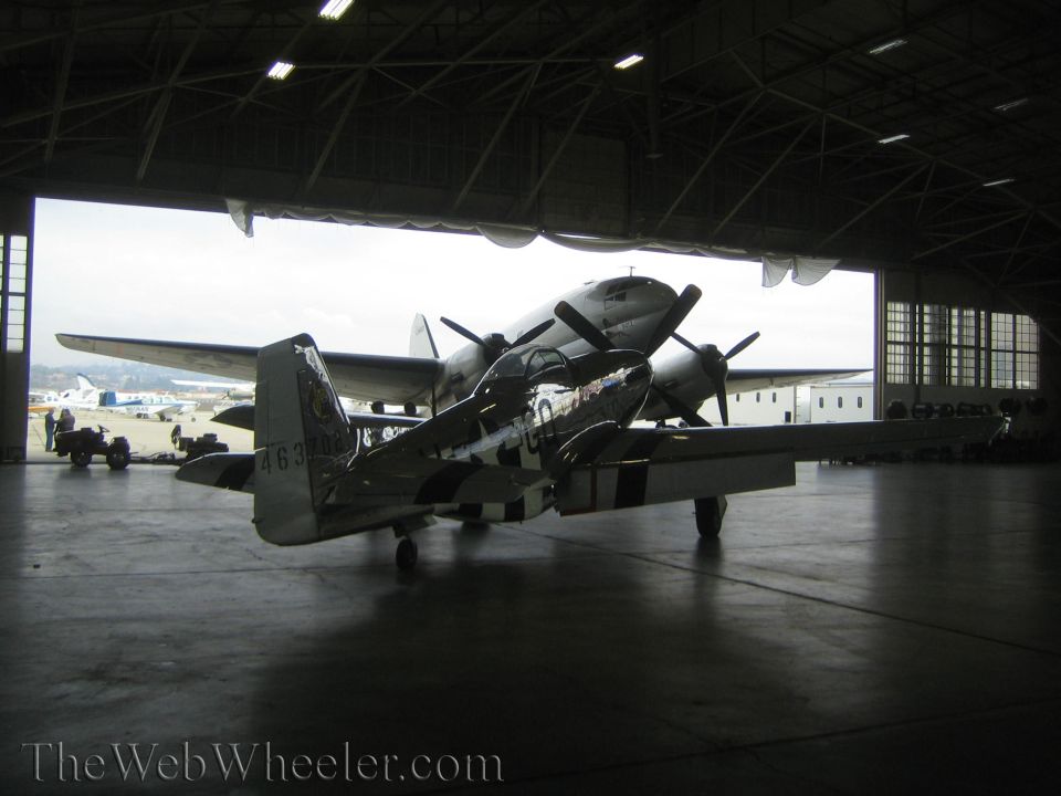 Silver fighter aircraft with invasion stripes parked under wing of larger plane in hangar