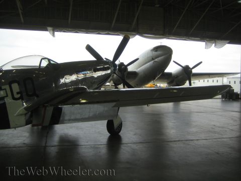 1990 Ford Bronco - polished silver fighter aircraft marked 'GQ' sits under the wing of a multi-engine plane inside a dim hangar.